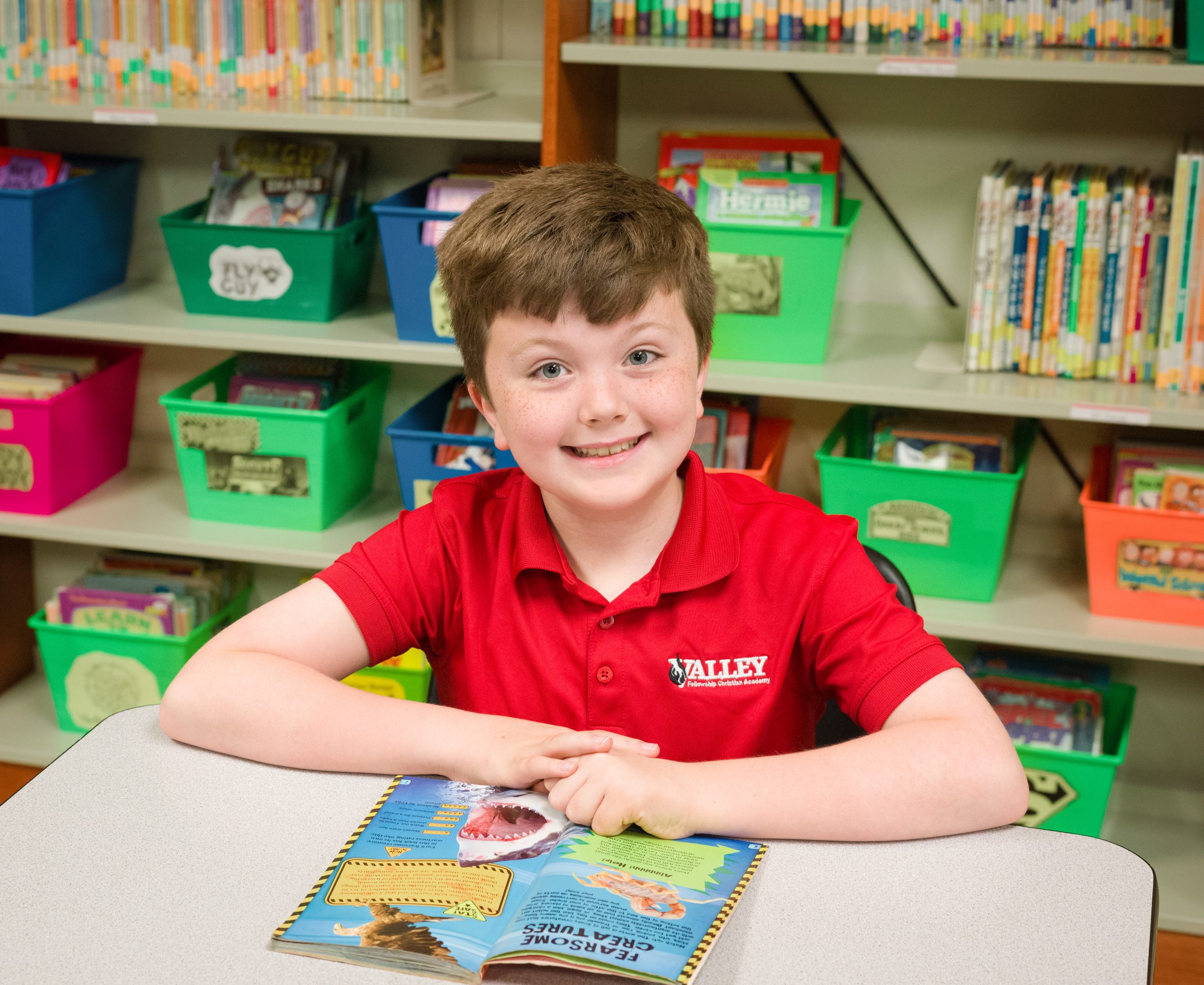 Little boy in a red VFCA T-shirt smiling while reading a book